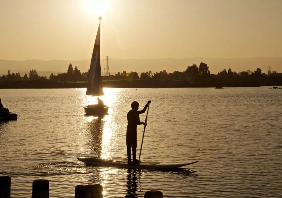 Paddle boarder with sailboat in background at sunset