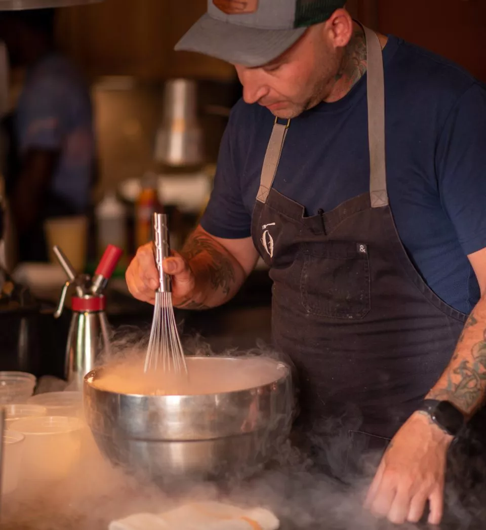 Chef stirring a stainless steel bowl with a whisk with fog billowing out