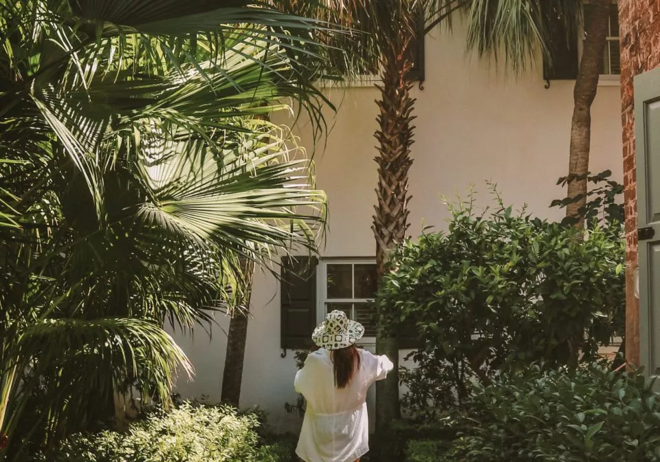 Wide view of the back of a woman in white shirt and hat surrounded by bushes and palm trees